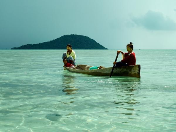 Two young women in a wooden canoe on jade green sea water with a small island on the background.
