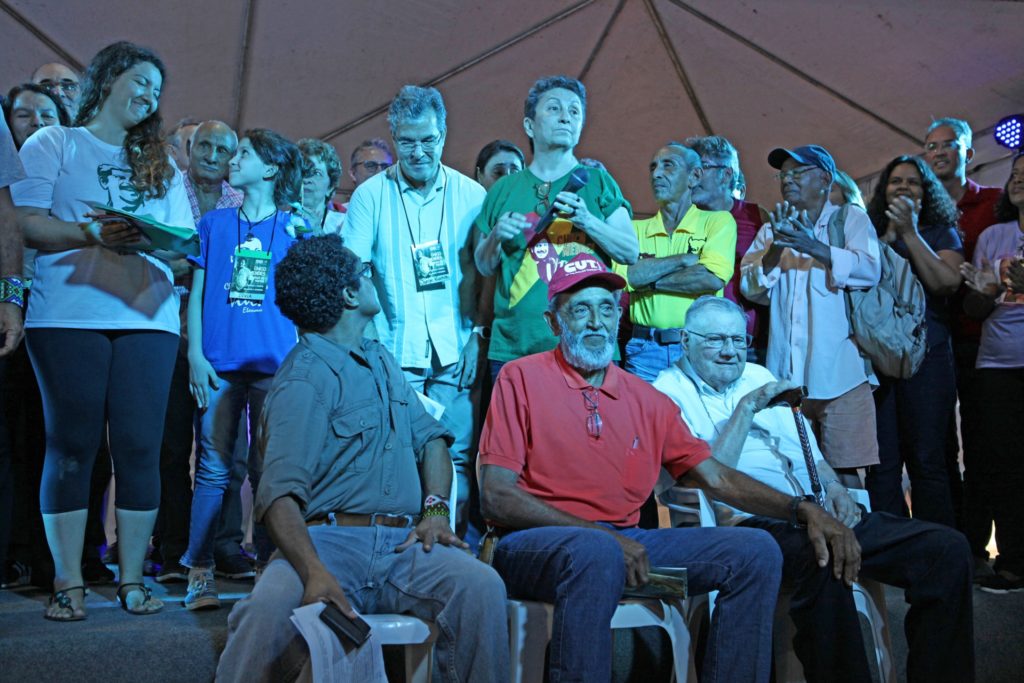 Miehiä ja naisia istumassa ja seisomassa suuren teltan esiintymislavalla.