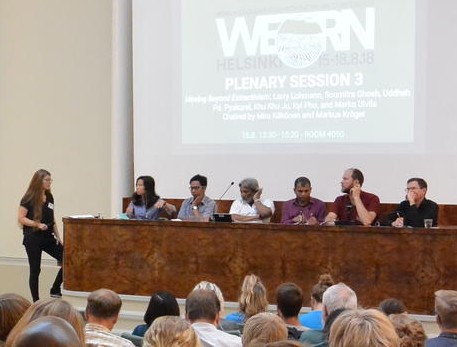 Six conference panelists sitting in front of audience.