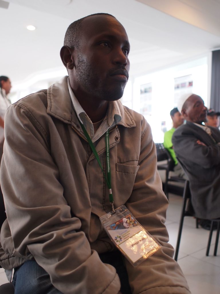 Man sits indoors wearing jacket and a conference badge.