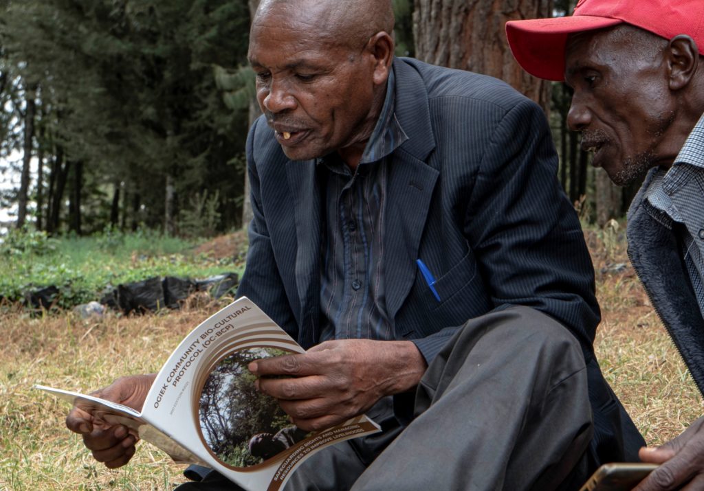 Two elderly black men sitting outdoors near forest and reading a booklet.